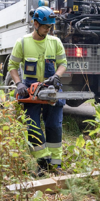 UVR:n työntekijä turvavausteissaan metsässä saha kädessään. Taustalla kuorma-auto