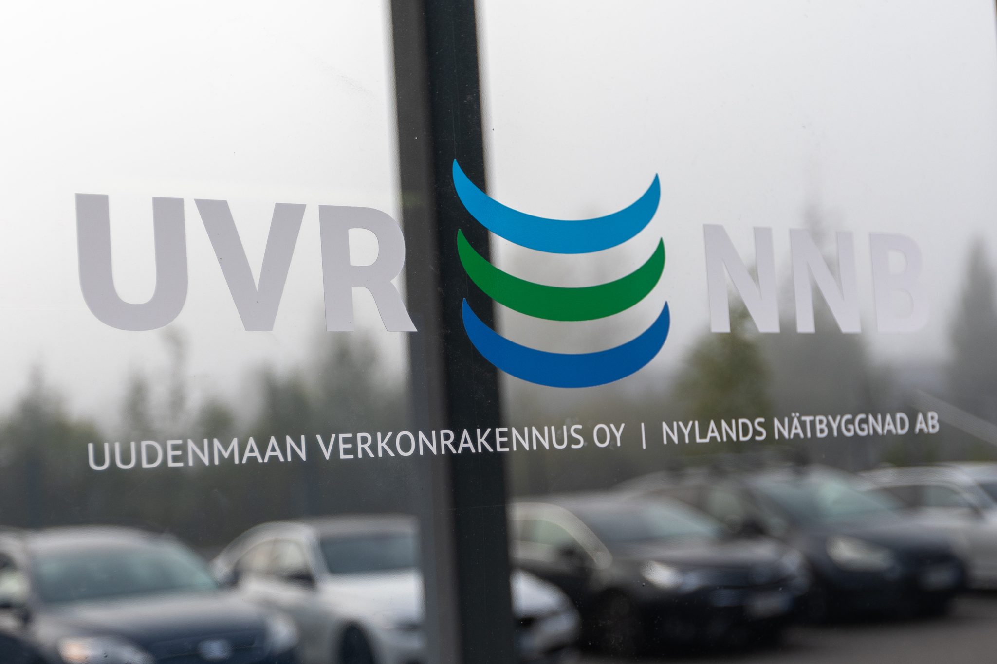 UVR:n toimitalon ikkunasta näkymä ulos, taustalla autoja. Ikkunassa näkyy UVR – NNB -logoteippaus Otsikko: Uudenmaan verkonrakennus Oy, Nylands nätbyggnad Ab